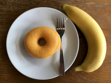 one fresh round donut with steel fork in white round ceramic plate and one fresh banana ready to eat and enjoy on natural wood backgroundの写真素材
