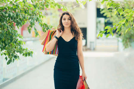 Cheerful lady in stylish black dress carrying on colorful shopping bags while walking at store. Attractive brunette looks satisfied after shopping at mall.の写真素材