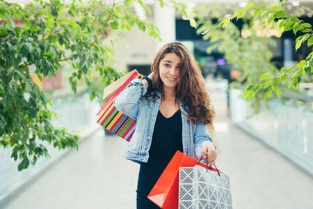Cheerful lady in stylish black dress carrying on colorful shopping bags while walking at store. Attractive brunette looks satisfied after shopping at mall.の写真素材