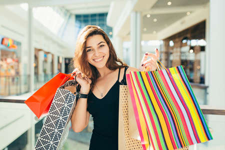 Cheerful lady in stylish black dress carrying on colorful shopping bags while walking at store. Attractive brunette looks satisfied after shopping at mall.の写真素材
