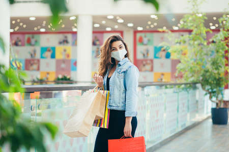 Happy young woman in medical mask walking at mall with lots of shopping bags in hands. Concept of healthcare and prevention from coronavirus.の写真素材