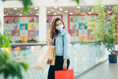 Happy young woman in medical mask walking at mall with lots of shopping bags in hands. Concept of healthcare and prevention from coronavirus.の写真素材