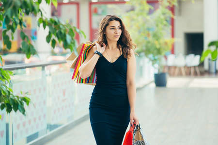 Cheerful lady in stylish black dress carrying on colorful shopping bags while walking at store. Attractive brunette looks satisfied after shopping at mall.の写真素材