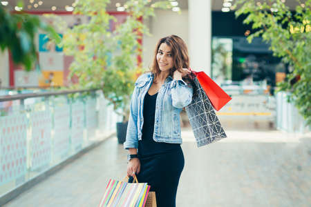 Cheerful lady in stylish black dress carrying on colorful shopping bags while walking at store. Attractive brunette looks satisfied after shopping at mall.の写真素材