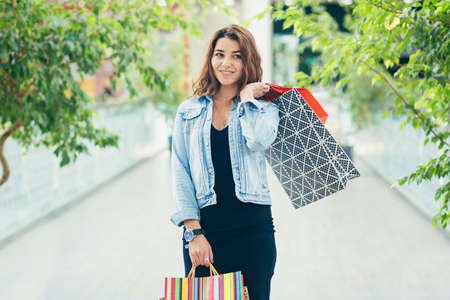 Cheerful lady in stylish black dress carrying on colorful shopping bags while walking at store. Attractive brunette looks satisfied after shopping at mall.の写真素材