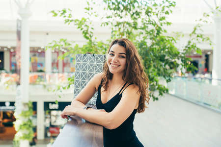 Cheerful lady in stylish black dress carrying on colorful shopping bags while walking at store. Attractive brunette looks satisfied after shopping at mall.の写真素材