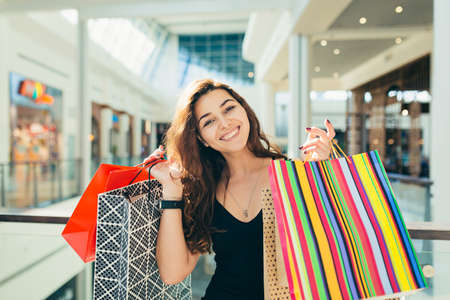 Cheerful lady in stylish black dress carrying on colorful shopping bags while walking at store. Attractive brunette looks satisfied after shopping at mall.の写真素材