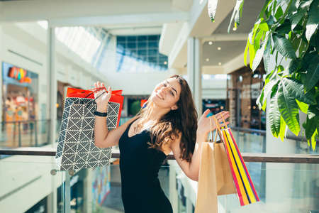 Cheerful lady in stylish black dress carrying on colorful shopping bags while walking at store. Attractive brunette looks satisfied after shopping at mall.の写真素材