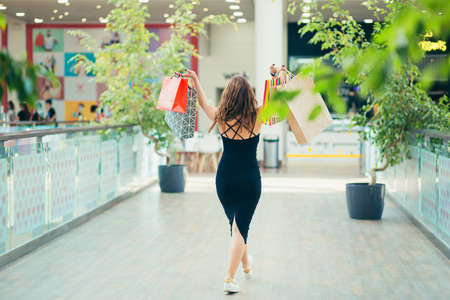 Cheerful lady in stylish black dress carrying on colorful shopping bags while walking at store. Attractive brunette looks satisfied after shopping at mall.の写真素材