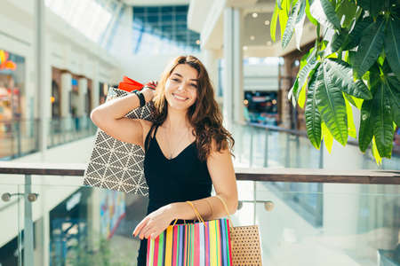 Cheerful lady in stylish black dress carrying on colorful shopping bags while walking at store. Attractive brunette looks satisfied after shopping at mall.の写真素材