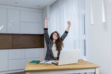 Young woman rejoices at her finished work with raised fists, sitting at her deskの写真素材