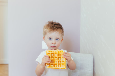 Little boy in a white bodysuit eating breakfast with a waffle in his kitchenの写真素材