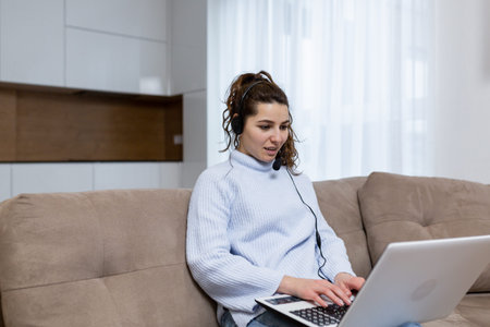 A young woman employee of a call center, provides online consultation staying at home, sitting on...