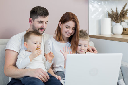 Young family, husband, wife and two young sons, sitting at home in the kitchen, and communicating via video link with family, using laptopの写真素材