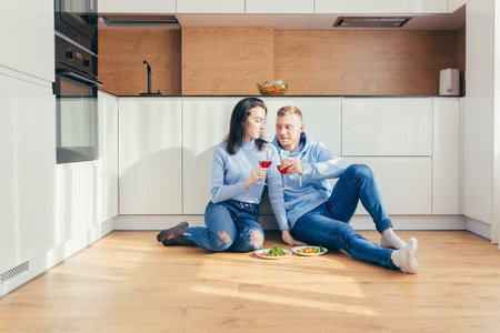 Couple in love eating a salad of vegetables, sitting on the floor in the kitchen, and drinking wineの写真素材