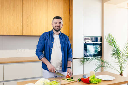 A young man spends a day at home, prepares a breakfast of vegetables in the kitchen, a man in home clothes and with a beard, uses a laptop to learn online cookingの写真素材
