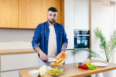 A young man spends the day at home, preparing breakfast in the kitchen with vegetables, a man in home clothes and with a beardの写真素材