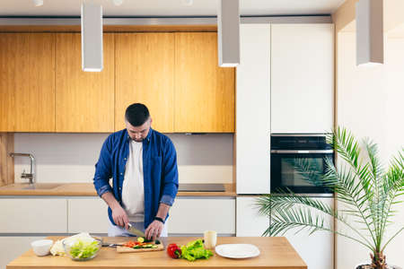 A young man spends the day at home, preparing breakfast in the kitchen with vegetables, a man in home clothes and with a beardの写真素材