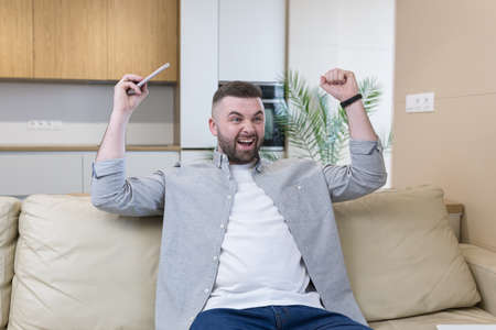 Smiling young man holding mobile phone while sitting on a sofa at home and celebratingの写真素材