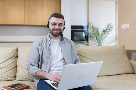 A man with a beard works from home sitting on the couch, uses a laptop and a headset with a microphone conducts an online consultationの写真素材