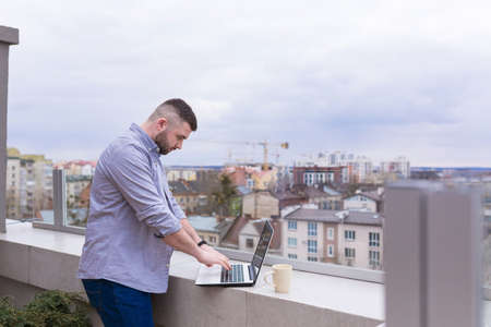 Male businessman, in casual clothes relaxing on the terrace overlooking the city, uses the phone, and uses a laptop while drinking coffeeの写真素材