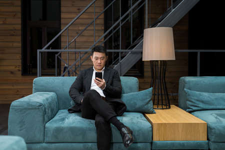 Successful Asian businessman manager, in a black business suit, uses the phone for video calls and conferences relaxing in a restaurant or hotel, and in a stylish office explainsの写真素材