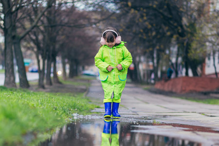 girl jumps on puddles in a green raincoat and blue boots, against the backdrop of the city and trees, in the spring after the rainの写真素材