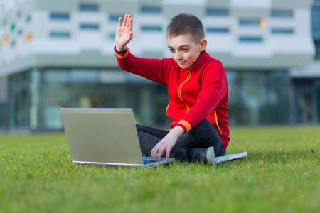 Little teenager boy, a student of a private school, in his free time communicates with his parents sitting on the lawn uses a laptopの写真素材