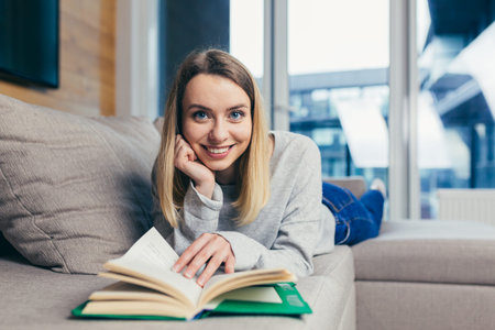 Young beautiful blonde woman reads an interesting book while lying on the couch, resting after work