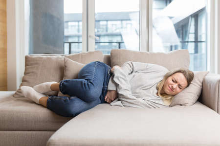 young woman sitting or lying on sofa or couch at home with severe abdominal pain. Female on bed holds her stomach with premenstrual, menstrual or poisoning. blonde girl suffers from abdominal crampsの写真素材