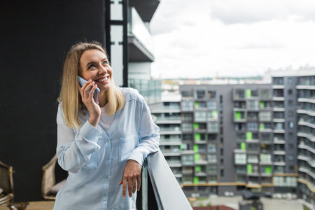 Beautiful blonde woman uses a mobile phone while standing on the balcony of an apartment building