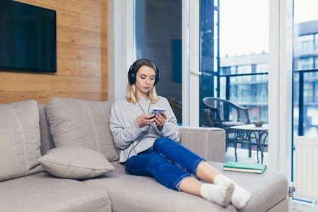 Young beautiful woman resting at home listening to audio podcast sitting on sofa, using headphones and smartphone, for online listeningの写真素材