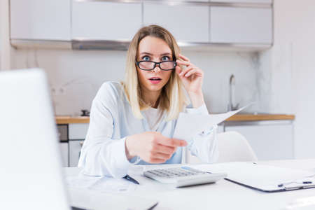 young female housewife sitting a table in house kitchen an incomprehensible deal with paperwork bills and loans. Woman is exhausted and frustrated with documents of enterprise or family budget at homeの写真素材