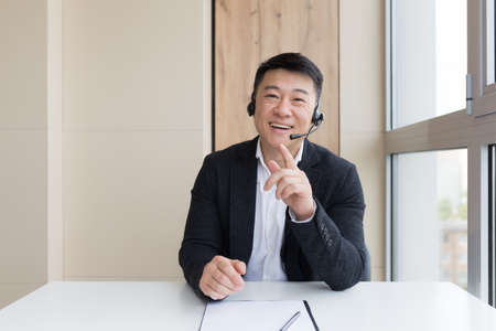 Male businessman conducts an online consultation explains cheerfully gestures with his hands using a headset, and online callの写真素材