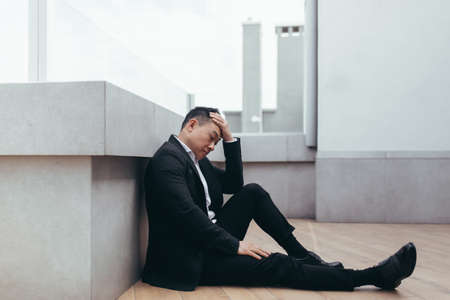 young asian depressed man sitting on outdoors street near office business center building. The upset male businessman lost his job due to the financial crisis and quarantine. Businessman has problemsの写真素材