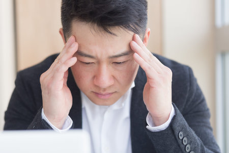 young asian man, office worker sitting at workplace holding hands massages forehead and head with...