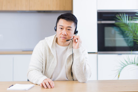 Asian man talking online on video call at home room with headset. Male in casual clothes looking at...
