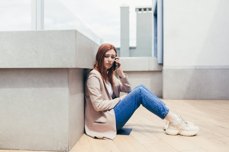 young frustrated woman sitting on sidewalk near a depressed business office center has lost job,...