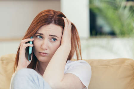 Young beautiful woman sitting alone at home on the couch, feeling pain and depression complains to a friend on the phoneの写真素材