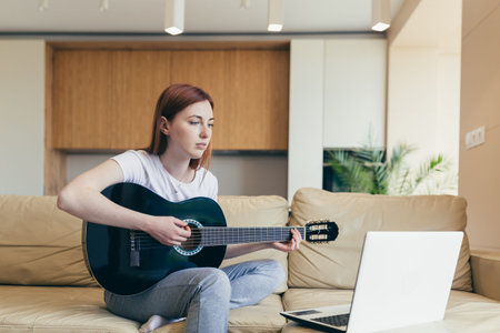 young red haired girl learns to play the guitar with the help of video tutorial lessons. female woman home at leisure sitting on sofa with laptop studying online musical instrument. Hobbies distance
