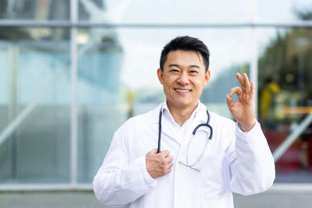 portrait of male cheerful asian doctor smiling and looking at camera showing ok hand sign on modern clinic outdoor backgroundの写真素材