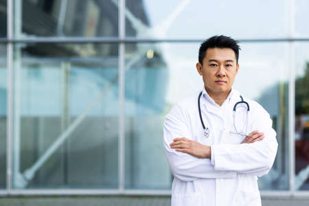 portrait of serious asian doctor with folded arms on background of modern clinic outdoorsの写真素材