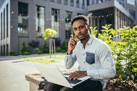 young african student studies online sitting on a bench near the university, uses a laptop and...