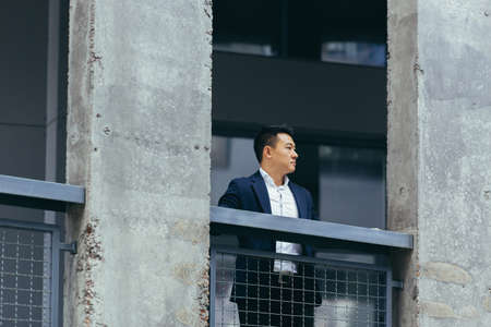 Young Asian businessman standing near modern black office building made of concrete, on terrace balcony with columnsの写真素材
