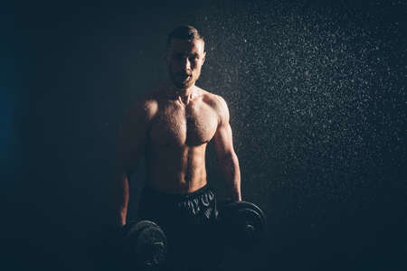 Male blond athlete lifts dumbbells with hand on biceps studio photo with raindropsの写真素材