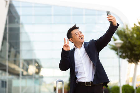 Asian tourist businessman arrived at a business conference takes photos near the airport and communicates by video with colleagues using a smartphoneの写真素材