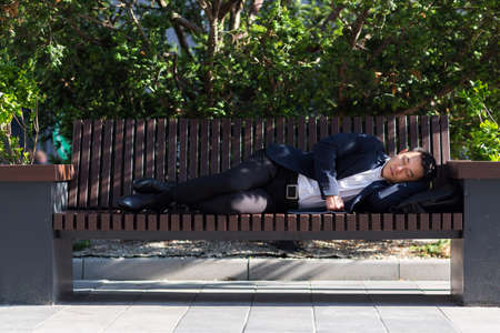 A tired Asian worker is lying in front of the company during a break from work. Rest after a hard day's work on a park benchの写真素材