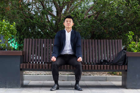 Asian businessman breathing exercises trying to calm stress, sitting on a bench during a lunch break in a business suitの写真素材