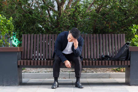 exhausted young asian business man office worker sitting on bench in city park outdoors. stressed overworked male with a headache massages head. Depressed Tired pain suffering pain sick workの写真素材