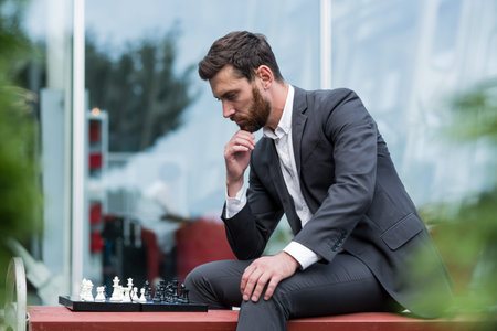 Banker male businessman playing chess sitting on bench near office, pensive making strategic move
